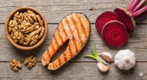 Image 2 - A vibrant flat-lay of heart-healthy foods including a grilled salmon fillet, a bowl of fresh walnuts, sliced red beets, and several cloves of garlic on a wooden cutting board.