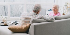 A senior couple in a bright living room, with one individual adjusting their hearing aid.