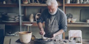 An elderly person crafting a clay bowl on a pottery wheel in a sunlit studio.