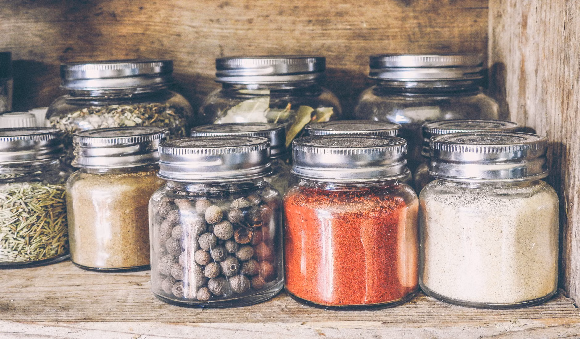Organized glass jars of dried herbs on a wooden pantry shelf.