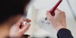 Close-up of hands sketching a plant in a sketchbook near a window.