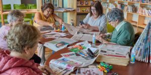 A group of older adults engaged in a friendly craft-sharing session in a sunlit community center.
