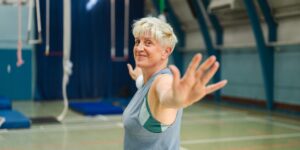 An older adult performing a seated arm stretch in a bright, comfortable living room.