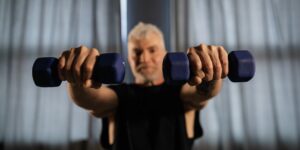 A senior person performing seated shoulder exercises in a sunlit room.