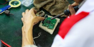 An older adult repairing an electronic assistive device at a desk.