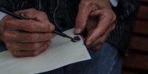 A close-up of an elderly person's hands practicing calligraphy in a sunlit room.