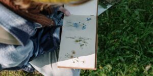 An older adult drawing a wildflower in a sketchbook with colored pencils in soft morning light.